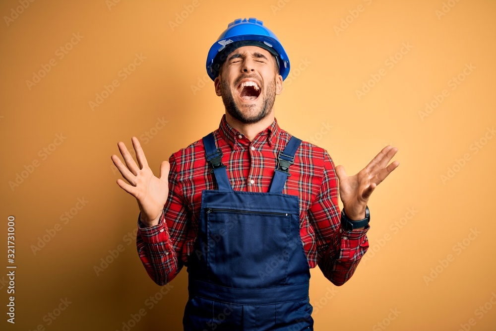 Fototapeta premium Young builder man wearing construction uniform and safety helmet over yellow background celebrating mad and crazy for success with arms raised and closed eyes screaming excited. Winner concept
