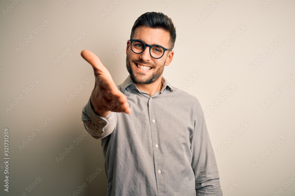 Young handsome man wearing elegant shirt and glasses over isolated white background smiling friendly offering handshake as greeting and welcoming. Successful business. Young handsome man wearing elegant shirt and glasses over isolated white background smiling friendly offering handshake as greeting and welcoming. Successful business.