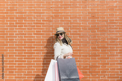Smiling attractive woman with hat and white coat, carries bags of gifts after shopping