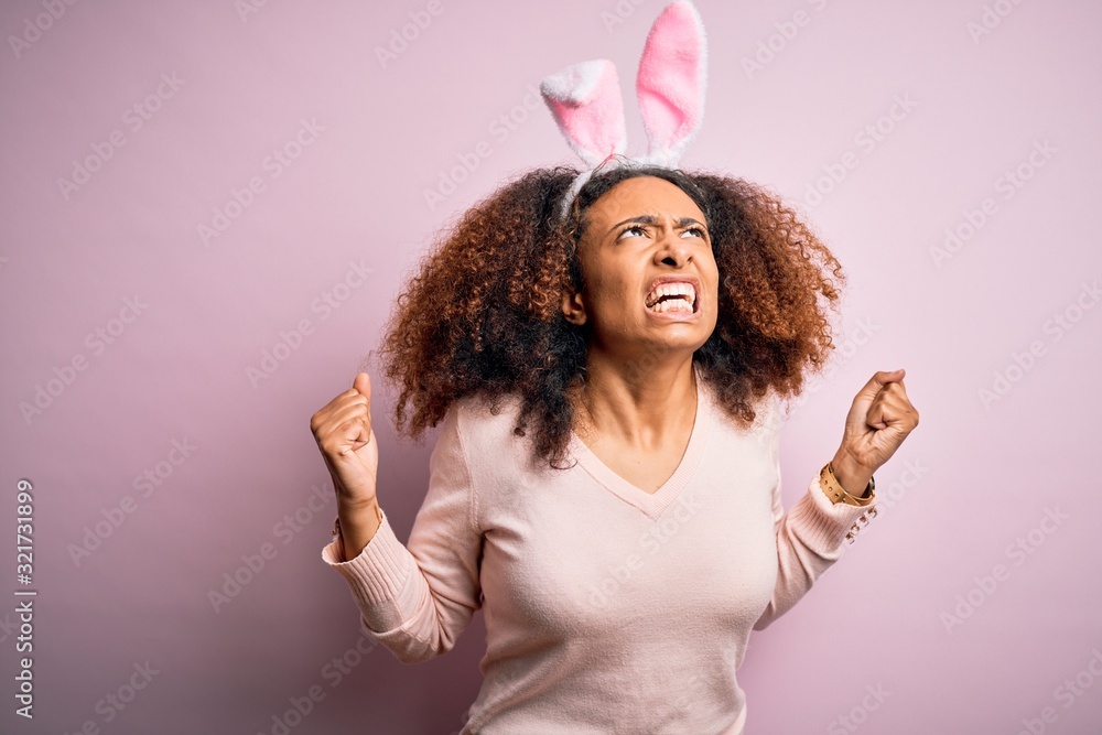 Young african american woman with afro hair wearing bunny ears over pink background crazy and mad shouting and yelling with aggressive expression and arms raised. Frustration concept. Young african american woman with afro hair wearing bunny ears over pink background crazy and mad shouting and yelling with aggressive expression and arms raised. Frustration concept.
