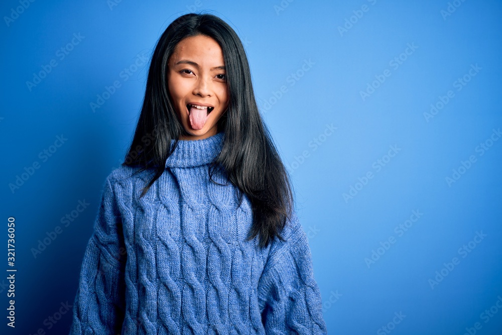 Young beautiful chinese woman wearing casual sweater over isolated blue background sticking tongue out happy with funny expression. Emotion concept. Young beautiful chinese woman wearing casual sweater over isolated blue background sticking tongue out happy with funny expression. Emotion concept.