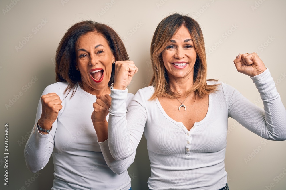Middle age beautiful couple of sisters wearing casual t-shirt over isolated white background celebrating surprised and amazed for success with arms raised and open eyes. Winner concept. Middle age beautiful couple of sisters wearing casual t-shirt over isolated white background celebrating surprised and amazed for success with arms raised and open eyes. Winner concept.