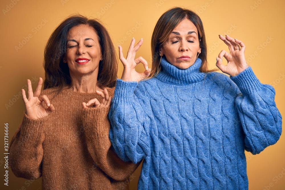 Middle age beautiful couple of sisters wearing casual sweater over isolated yellow background relax and smiling with eyes closed doing meditation gesture with fingers. Yoga concept. Middle age beautiful couple of sisters wearing casual sweater over isolated yellow background relax and smiling with eyes closed doing meditation gesture with fingers. Yoga concept.