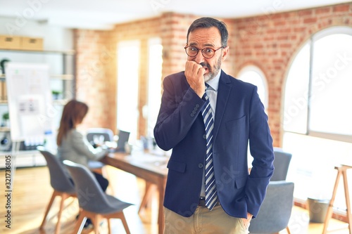 Middle age handsome businessman wearing glasses standing at the office looking stressed and nervous with hands on mouth biting nails. Anxiety problem.