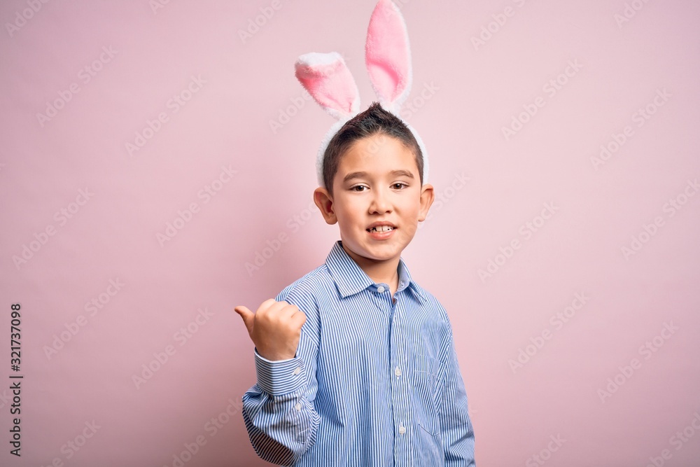 Young little boy kid wearing easter bunny ears over isolated pink background smiling with happy face looking and pointing to the side with thumb up. Young little boy kid wearing easter bunny ears over isolated pink background smiling with happy face looking and pointing to the side with thumb up.