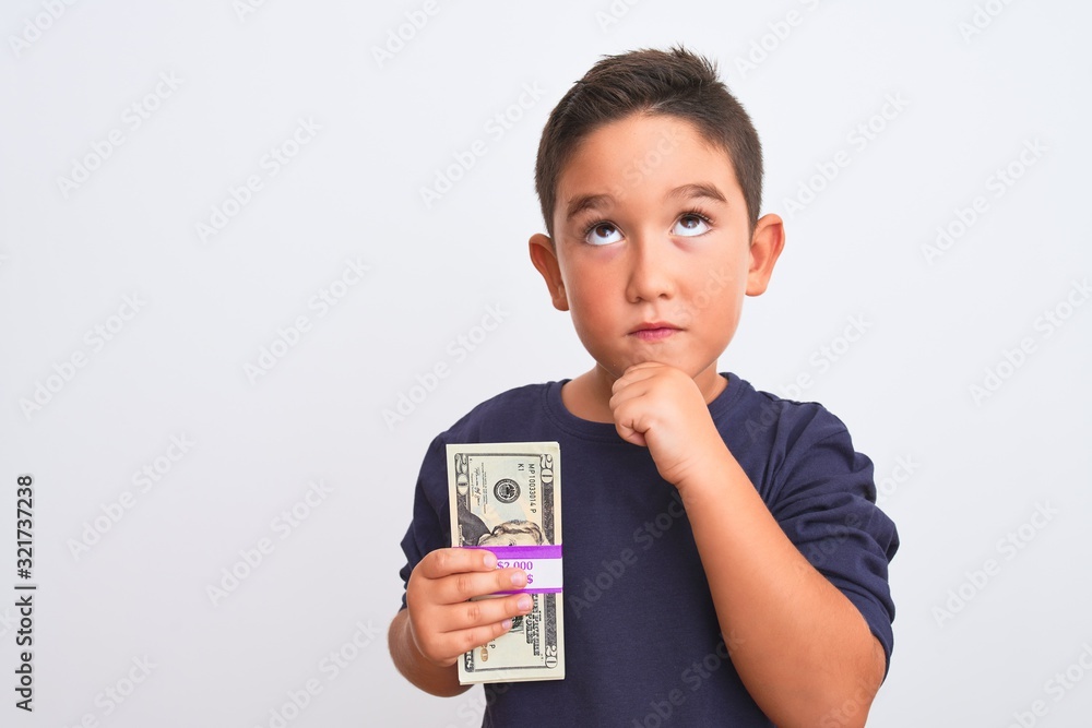 Beautiful kid boy holding dollars standing over isolated white background serious face thinking about question, very confused idea Beautiful kid boy holding dollars standing over isolated white background serious face thinking about question, very confused idea
