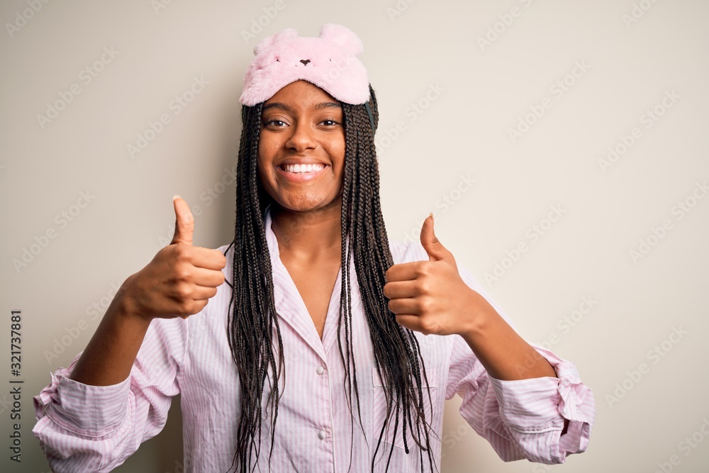 Young african american woman wearing pink pajama and sleep mask over isolated background success sign doing positive gesture with hand, thumbs up smiling and happy. Cheerful expression and winner Young african american woman wearing pink pajama and sleep mask over isolated background success sign doing positive gesture with hand, thumbs up smiling and happy. Cheerful expression and winner