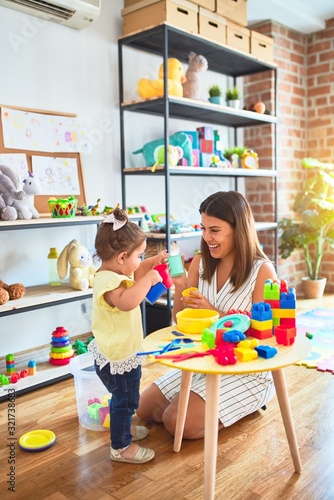 Young beautiful teacher and toddler playing with dishes, cutlery and cups toy on the table at kindergarten