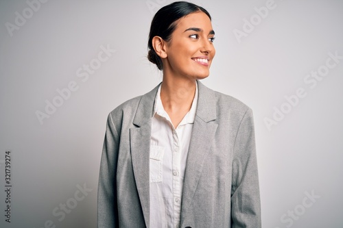 Fotografie Young beautiful brunette businesswoman wearing jacket standing over white background looking away to side with smile on face, natural expression