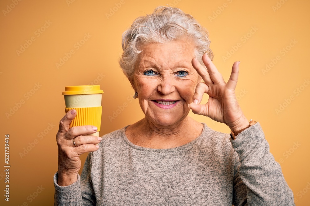 Senior beautiful woman drinking cup of coffee standing over isolated yellow background doing ok sign with fingers, excellent symbol Senior beautiful woman drinking cup of coffee standing over isolated yellow background doing ok sign with fingers, excellent symbol