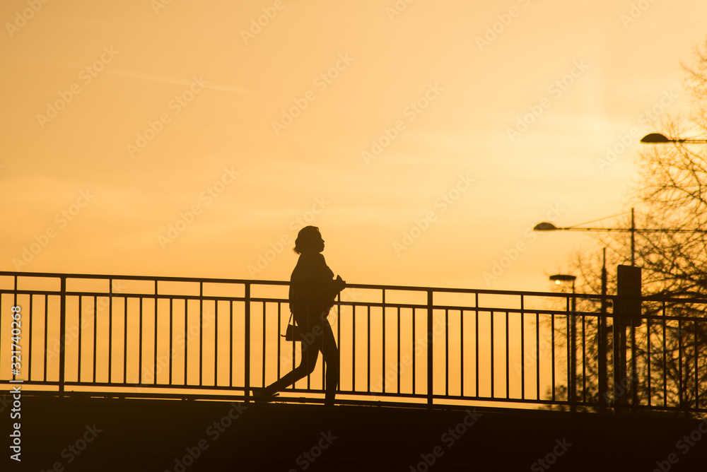 Silhouette of woman walking on bridge with smartphone in hands and earphones by sunset Silhouette of woman walking on bridge with smartphone in hands and earphones by sunset