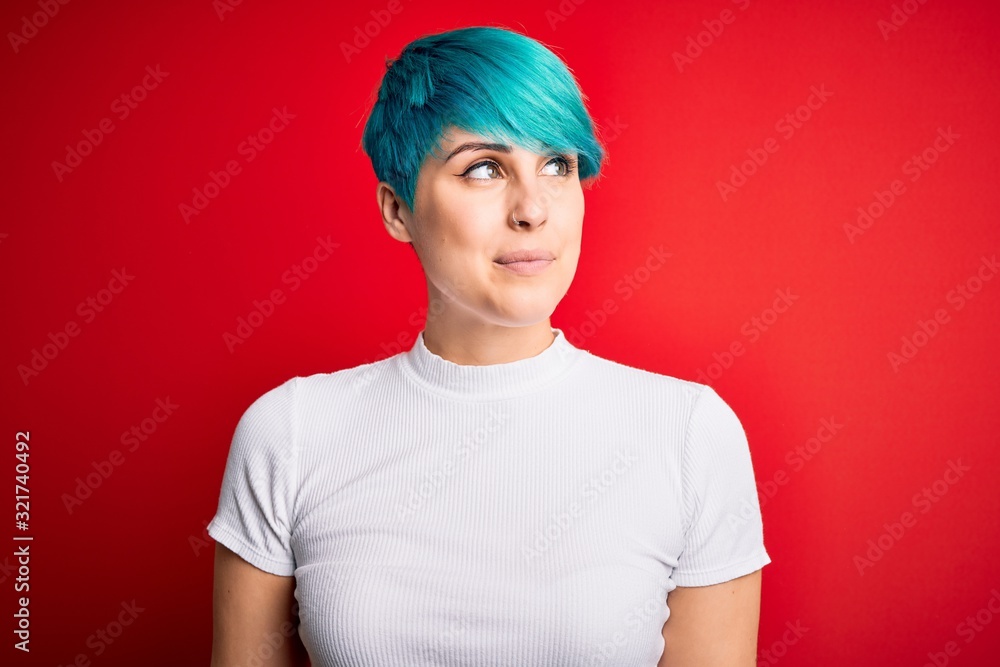 Fototapeta premium Young beautiful woman with blue fashion hair wearing casual t-shirt over red background smiling looking to the side and staring away thinking.