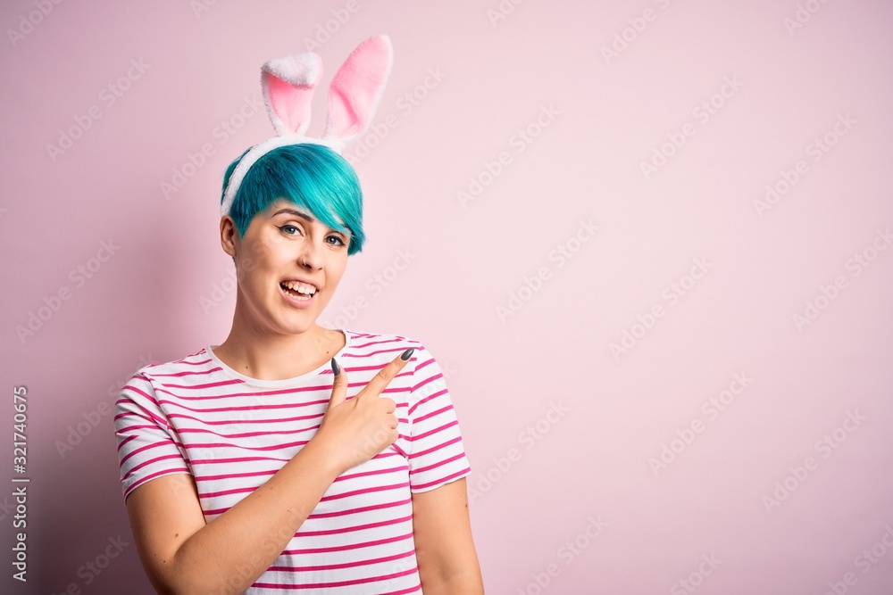 Young woman with fashion blue hair wearing easter rabbit ears over pink background cheerful with a smile of face pointing with hand and finger up to the side with happy and natural expression on face
