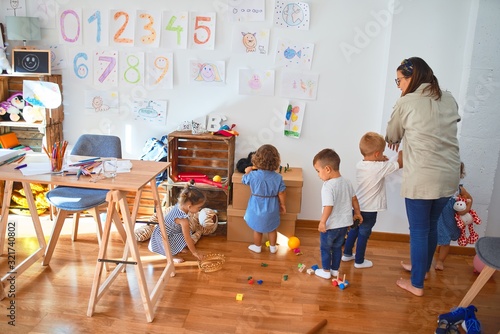 Fényképezés Beautiful teacher and group of toddlers playing around lots of toys at kindergar