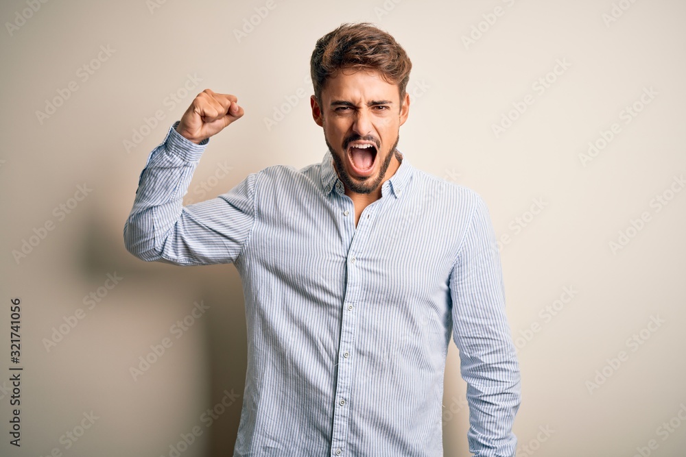Young handsome man with beard wearing striped shirt standing over white background angry and mad raising fist frustrated and furious while shouting with anger. Rage and aggressive concept. Young handsome man with beard wearing striped shirt standing over white background angry and mad raising fist frustrated and furious while shouting with anger. Rage and aggressive concept.