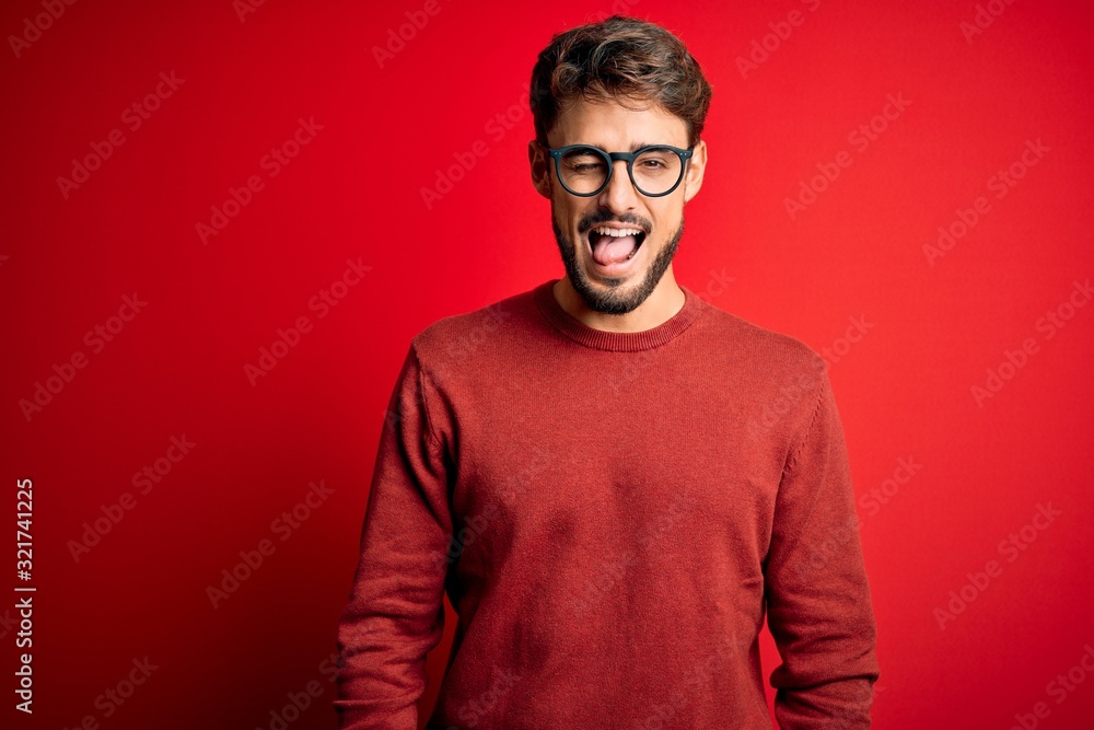 Young handsome man with beard wearing glasses and sweater standing over red background winking looking at the camera with sexy expression, cheerful and happy face.