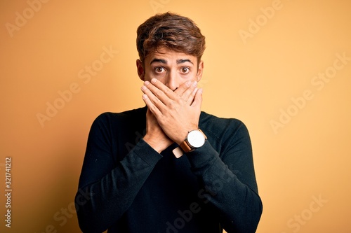 Young handsome man with beard wearing turtleneck sweater standing over yellow background shocked covering mouth with hands for mistake. Secret concept.