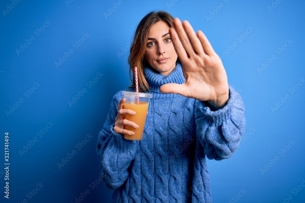 Young beautiful brunette woman drinking healthy orange juice over blue background with open hand doing stop sign with serious and confident expression, defense gesture Young beautiful brunette woman drinking healthy orange juice over blue background with open hand doing stop sign with serious and confident expression, defense gesture