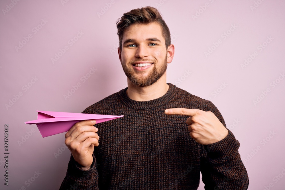 Young blond man with beard and blue eyes holding pink paper plane over isolated background very happy pointing with hand and finger Young blond man with beard and blue eyes holding pink paper plane over isolated background very happy pointing with hand and finger