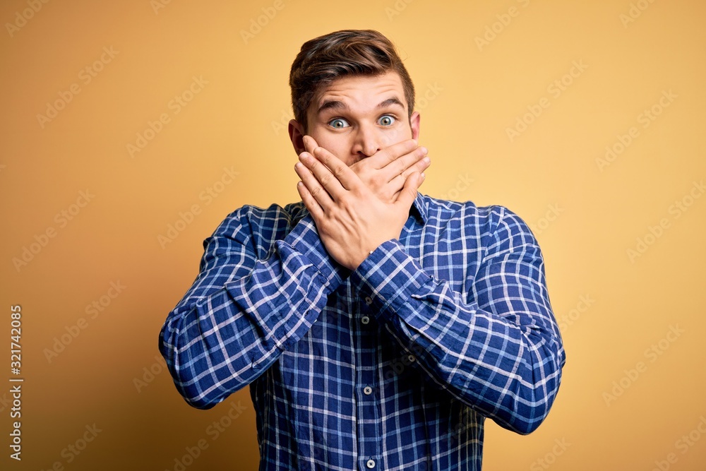 Young blond businessman with beard and blue eyes wearing shirt over yellow background shocked covering mouth with hands for mistake. Secret concept. Young blond businessman with beard and blue eyes wearing shirt over yellow background shocked covering mouth with hands for mistake. Secret concept.