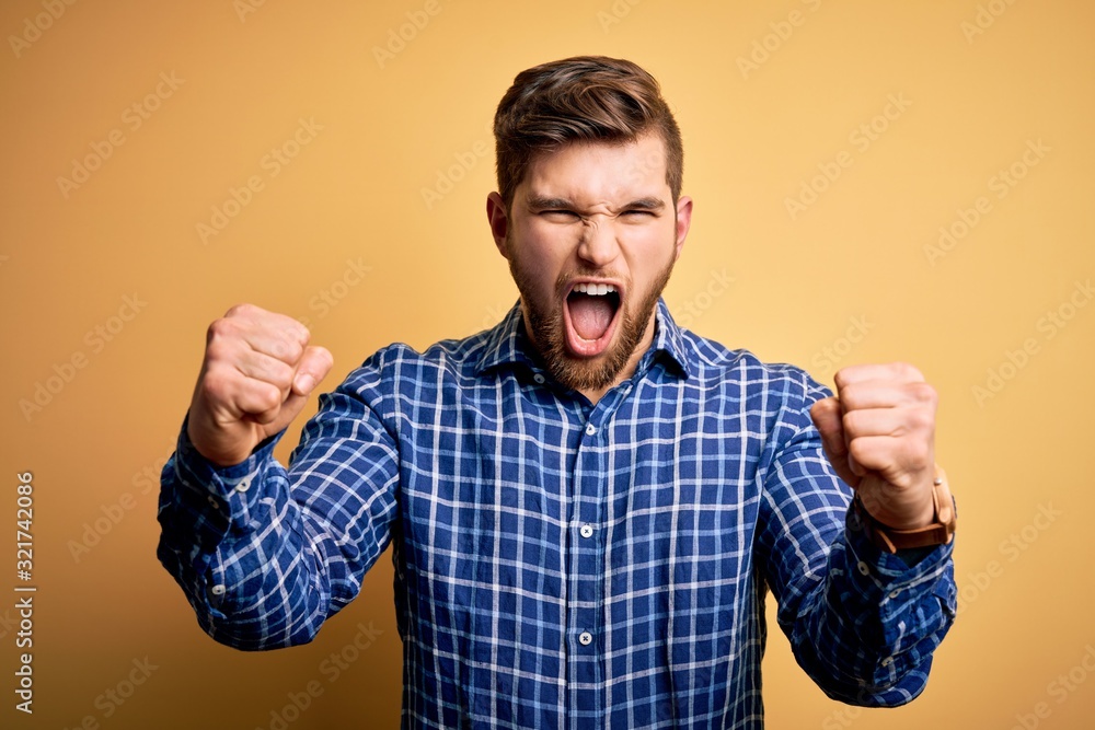 Young blond businessman with beard and blue eyes wearing shirt over yellow background angry and mad raising fists frustrated and furious while shouting with anger. Rage and aggressive concept. Young blond businessman with beard and blue eyes wearing shirt over yellow background angry and mad raising fists frustrated and furious while shouting with anger. Rage and aggressive concept.