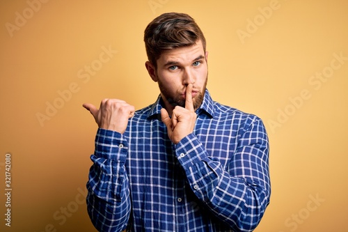 Young blond businessman with beard and blue eyes wearing shirt over yellow background asking to be quiet with finger on lips pointing with hand to the side. Silence and secret concept.
