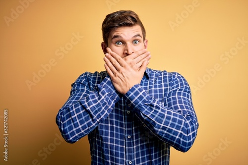 Young blond businessman with beard and blue eyes wearing shirt over yellow background shocked covering mouth with hands for mistake. Secret concept. © Krakenimages.com