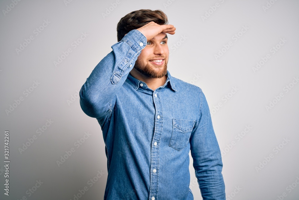 Young handsome blond man with beard and blue eyes wearing casual denim shirt very happy and smiling looking far away with hand over head. Searching concept. Young handsome blond man with beard and blue eyes wearing casual denim shirt very happy and smiling looking far away with hand over head. Searching concept.