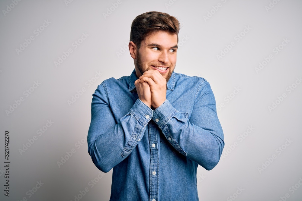 Young handsome blond man with beard and blue eyes wearing casual denim shirt laughing nervous and excited with hands on chin looking to the side Young handsome blond man with beard and blue eyes wearing casual denim shirt laughing nervous and excited with hands on chin looking to the side
