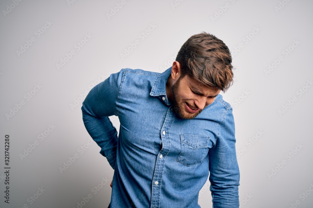 Young handsome blond man with beard and blue eyes wearing casual denim shirt Suffering of backache, touching back with hand, muscular pain Young handsome blond man with beard and blue eyes wearing casual denim shirt Suffering of backache, touching back with hand, muscular pain