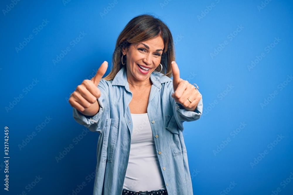 Middle age beautiful woman wearing casual shirt standing over isolated blue background approving doing positive gesture with hand, thumbs up smiling and happy for success. Winner gesture. Middle age beautiful woman wearing casual shirt standing over isolated blue background approving doing positive gesture with hand, thumbs up smiling and happy for success. Winner gesture.