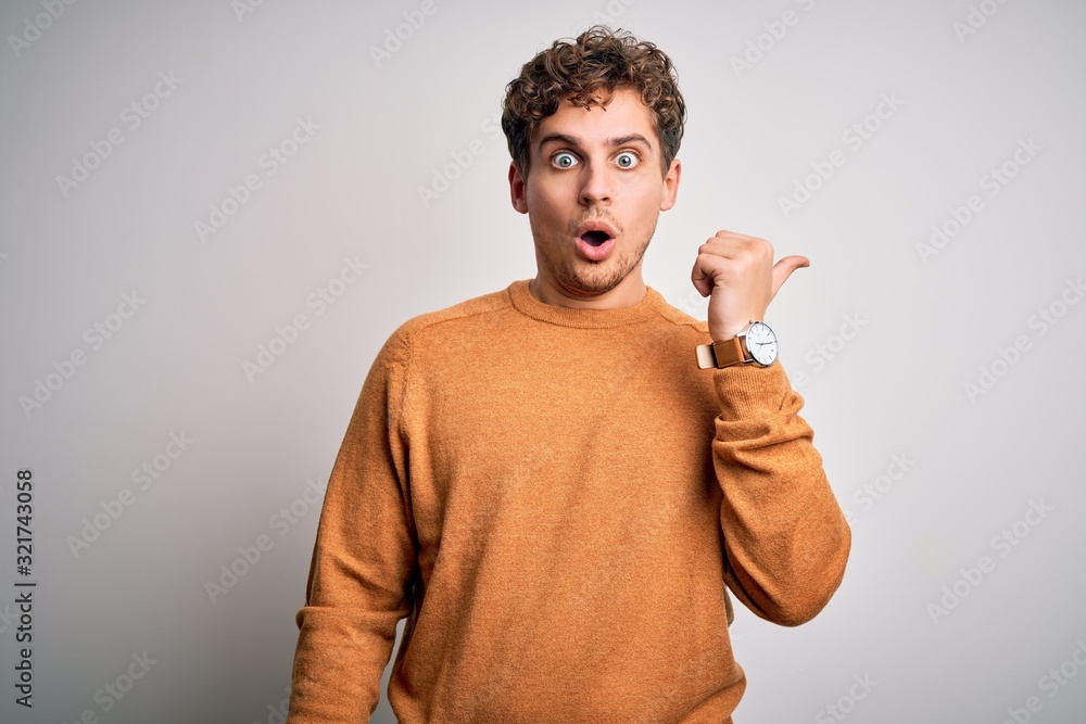 Young blond handsome man with curly hair wearing casual sweater over white background Surprised pointing with hand finger to the side, open mouth amazed expression. Young blond handsome man with curly hair wearing casual sweater over white background Surprised pointing with hand finger to the side, open mouth amazed expression.