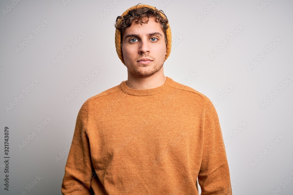 Young blond handsome man with curly hair wearing casual sweater and wool hat with serious expression on face. Simple and natural looking at the camera. Young blond handsome man with curly hair wearing casual sweater and wool hat with serious expression on face. Simple and natural looking at the camera.