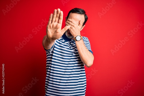 Middle age hoary man wearing casual striped polo standing over isolated red background covering eyes with hands and doing stop gesture with sad and fear expression. Embarrassed and negative concept.