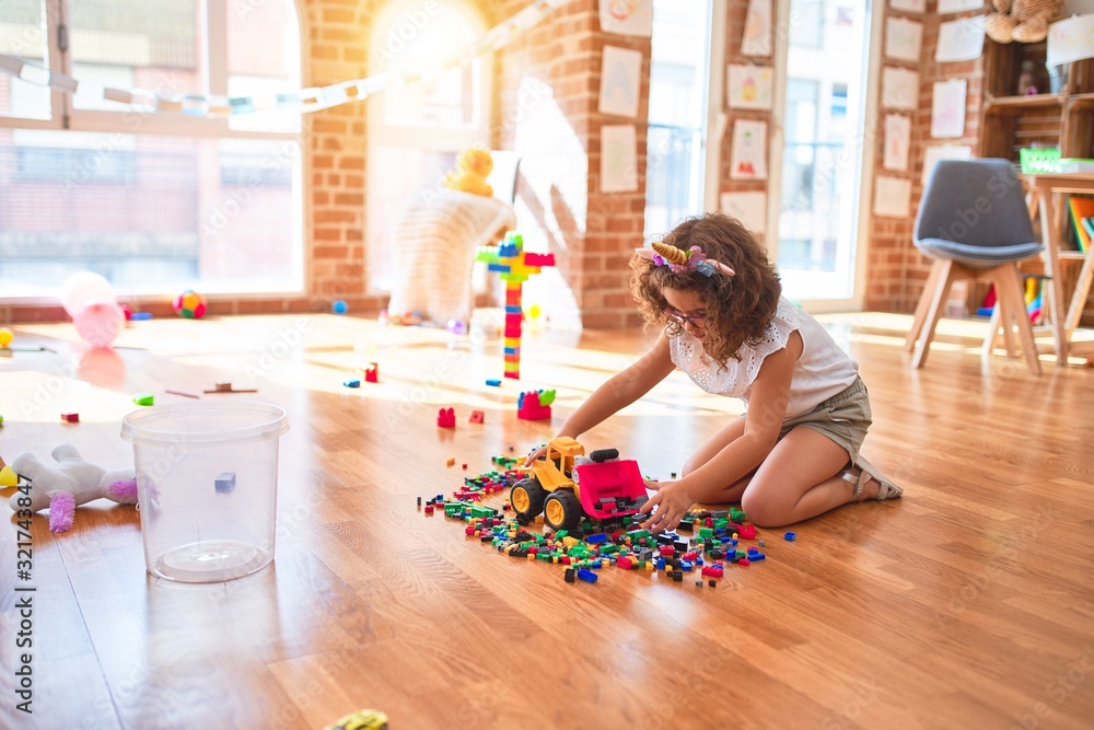 Beautiful toddler wearing glasses and unicorn diadem playing with tractor and building blocks at kindergarten - obrazy, fototapety, plakaty 