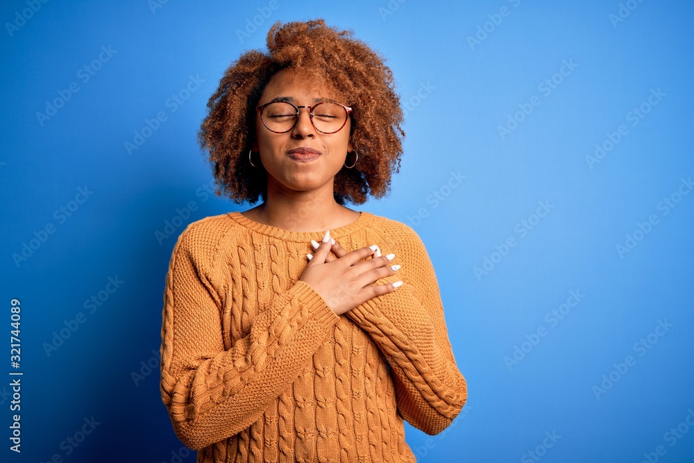 Young beautiful African American afro woman with curly hair wearing casual sweater smiling with hands on chest with closed eyes and grateful gesture on face. Health concept. Young beautiful African American afro woman with curly hair wearing casual sweater smiling with hands on chest with closed eyes and grateful gesture on face. Health concept.