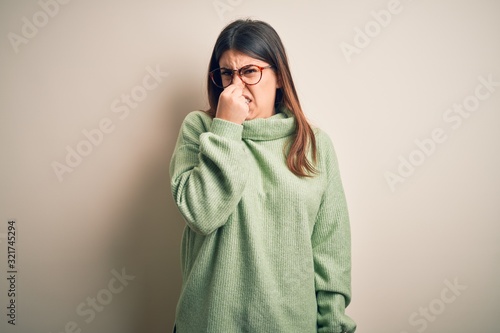Young beautiful woman wearing casual sweater standing over isolated white background smelling something stinky and disgusting, intolerable smell, holding breath with fingers on nose. Bad smell © Krakenimages.com