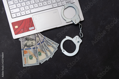 Handcuffs, laptop, money and credit card on dark background top view. Piracy, internet hacking and punishment for cybercrime concept.