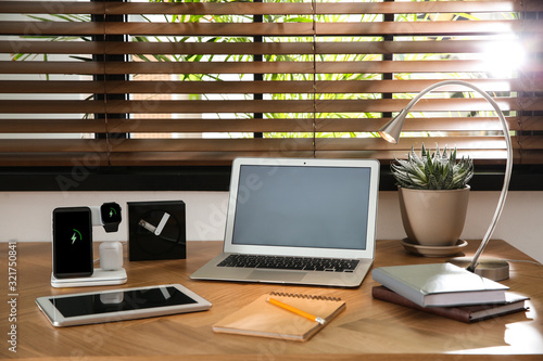 Mobile phone, earphones and smartwatch charging with wireless pad on wooden desk. Modern workplace device