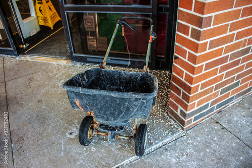 grungy two wheeled plastic salt spreader parked outside a store sliding glass door with salt scattered on the sidewalk around it after a snow photo