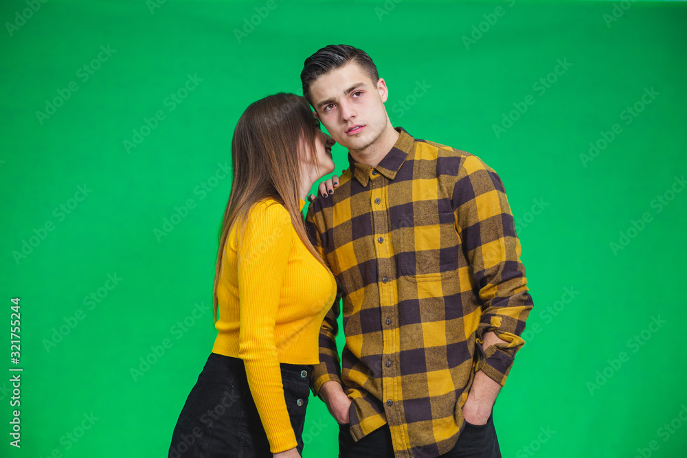 Young women sharing gossip and whispering secrets to her boyfriend, he is looking with puzzled face expression, standing over green background. Young women sharing gossip and whispering secrets to her boyfriend, he is looking with puzzled face expression, standing over green background.