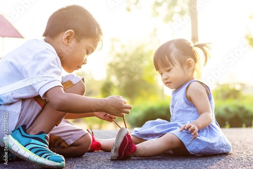 Soft focus. A young Asian brother help his little sister to tie her shoelaces. At the garden park in sunshine day summer season. Love and family concept.