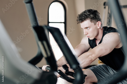 Concentrated serious fit young man riding bicycle simulator in gym