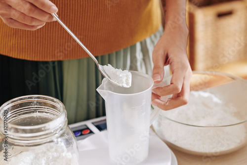 Close-up image of woman putting spoon of lye in plastic jar when making soap at home photo