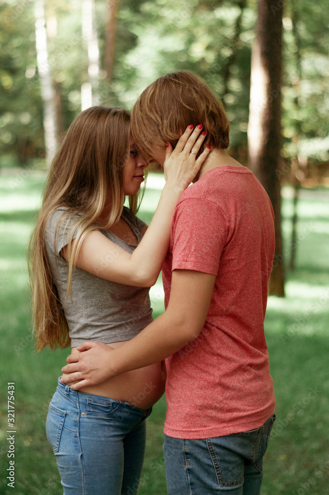 Fototapeta premium A happy married couple Husband hugs a beautiful adult pregnant wife. walks in a park. Second trimester. Holds his hands on her stomach. The benefits of walking during pregnancy, health care. Smiling