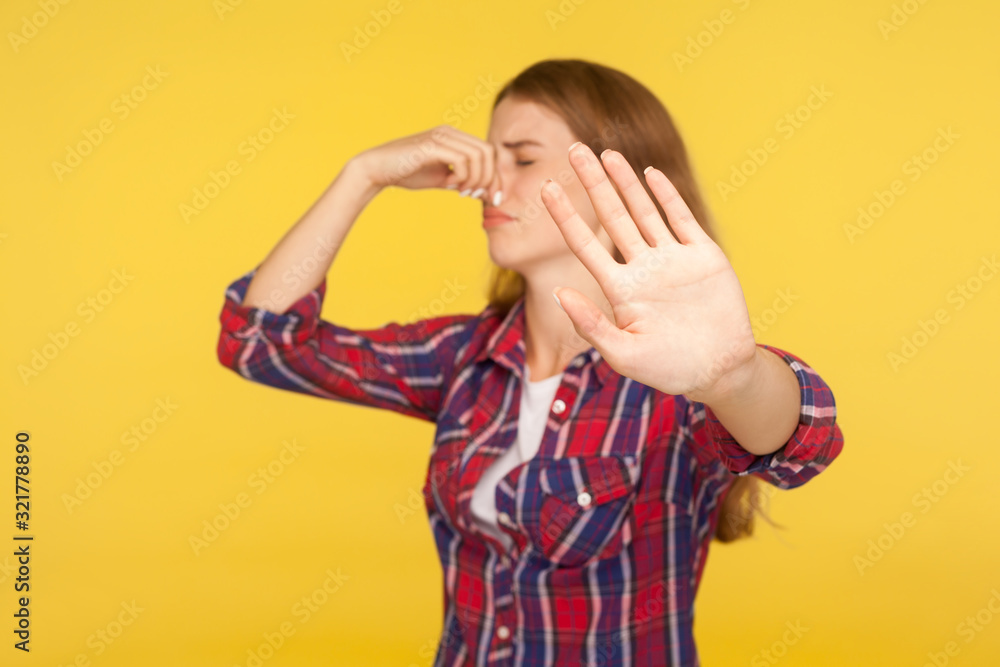 Bad smell. Portrait of displeased ginger girl in checkered shirt pinching nose and showing stop gesture, grimacing in disgust, avoiding stinky odor. indoor studio shot isolated on yellow background Bad smell. Portrait of displeased ginger girl in checkered shirt pinching nose and showing stop gesture, grimacing in disgust, avoiding stinky odor. indoor studio shot isolated on yellow background
