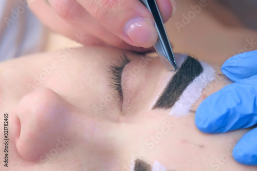 Professional beautician plucking eyebrows with tweezers to woman in beauty salon during tint eyebrow procedure, closeup view. Girl lying with closed eyes and brown natural henna on brows.