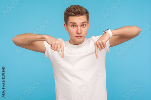 Come to me here now! Portrait of bossy man in white t-shirt pointing fingers down, demanding approach to him immediately, having control over situation. indoor studio shot isolated on blue background