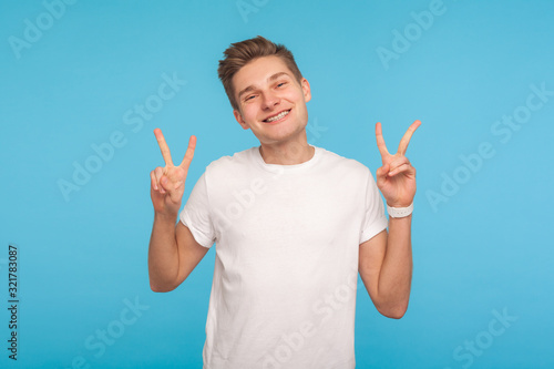 Portrait of happy delighted man in white t-shirt showing victory sign with double fingers, gesturing peace and looking at camera with friendly smile. indoor studio shot isolated on blue background