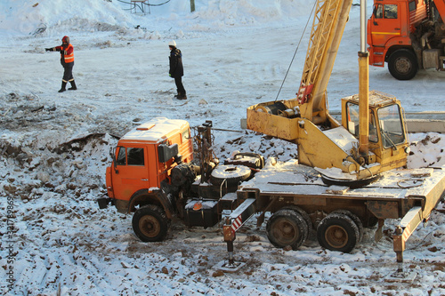 Truck crane truck with orange and yellow cabs with extended supports works at a construction site against the background of construction workers and snow in winter in Russia